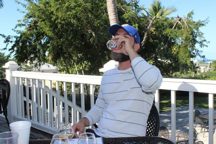 A person sitting at an outdoor table with a drink, surrounded by chairs and a white fence. 4
