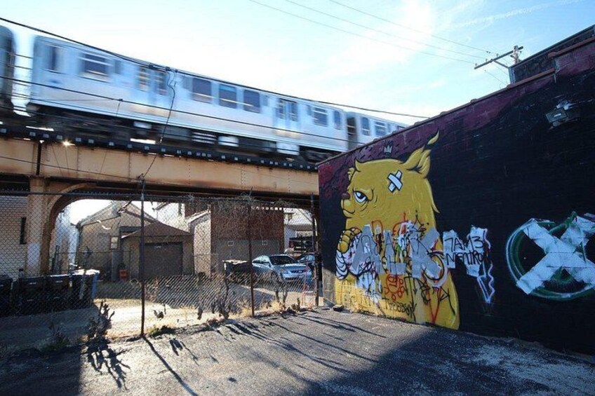 A train passing over an elevated bridge, a mural of a fox on a wall, and a fenced area with parked cars. 2