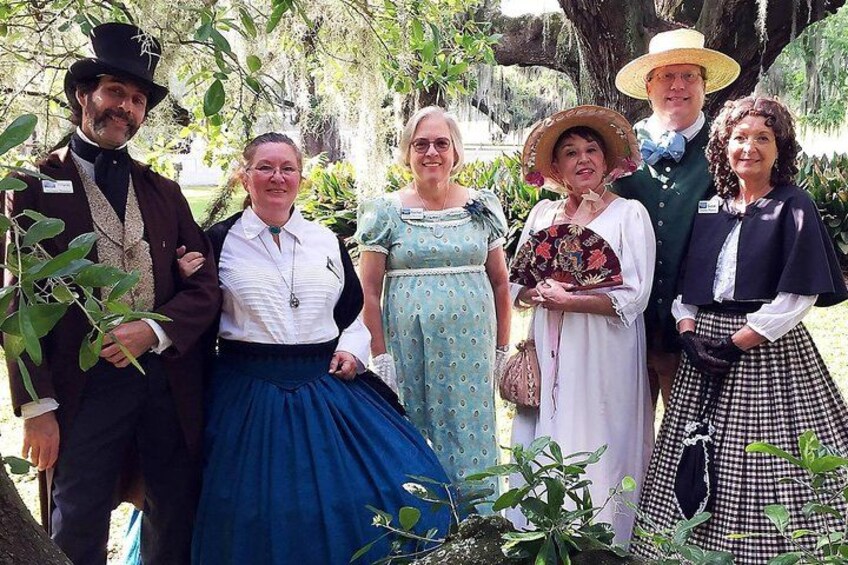 Un groupe de personnes vêtues de costumes d’époque se tient dans un jardin, entouré d’arbres et de plantes. 2