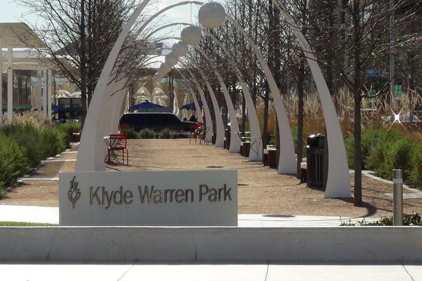 Ein Schild für den Klyde Warren Park, mit einem von weißen Bögen und Bäumen gesäumten Pfad. 3