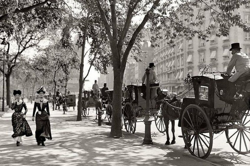 A historical street scene with horse-drawn carriages, pedestrians, and trees lining the road. 3