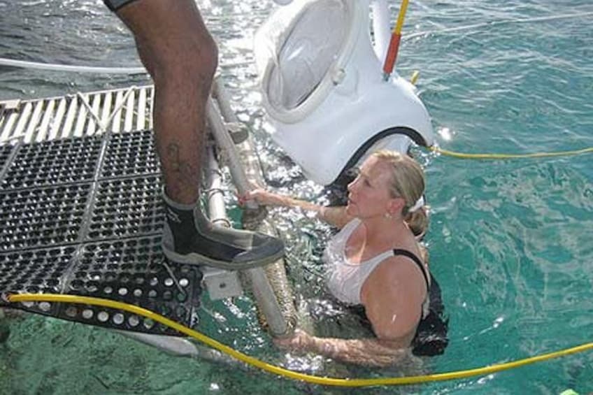 A person in a white tank top and black shorts swimming in clear blue water, holding onto a metal ladder. 5