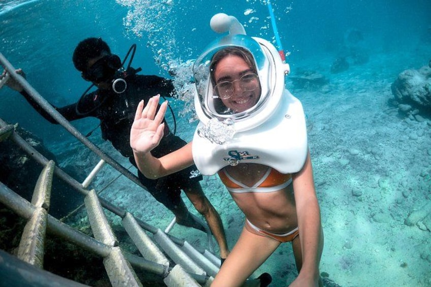 A person in a white scuba diving suit and orange bikini bottom descending a metal ladder underwater. 1