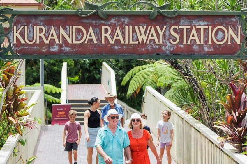 A sign reading 'Kuranda Railway Station' with people walking on a bridge. 5