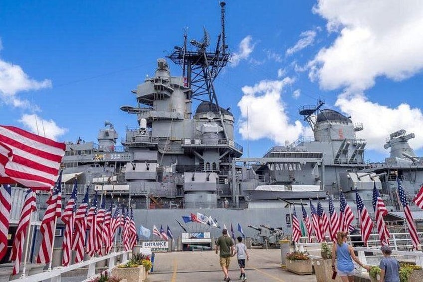A large naval ship with multiple American flags, a clear sky, and a few people walking around. 1