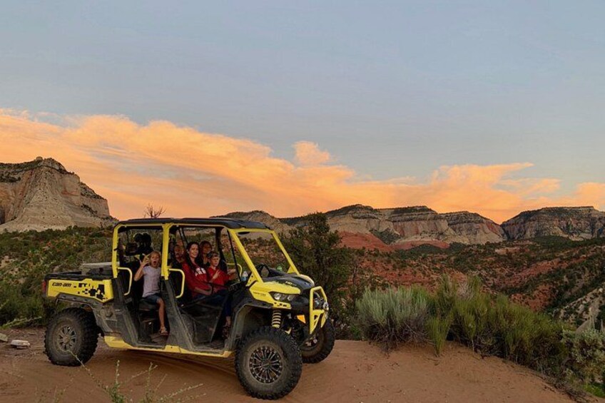 A yellow off-road vehicle with passengers, parked on a dirt path with a mountainous landscape in the background. 3