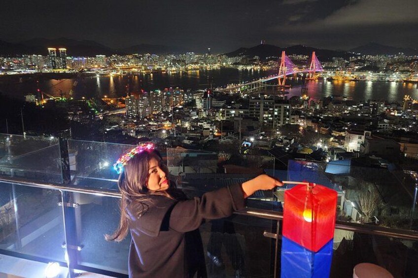 A person standing on a balcony overlooking a cityscape at night with a bridge in the background. 1