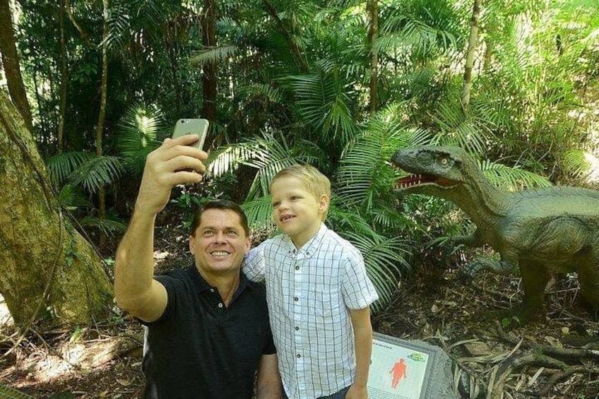 Two people taking a selfie with a dinosaur statue in a forested area. 3