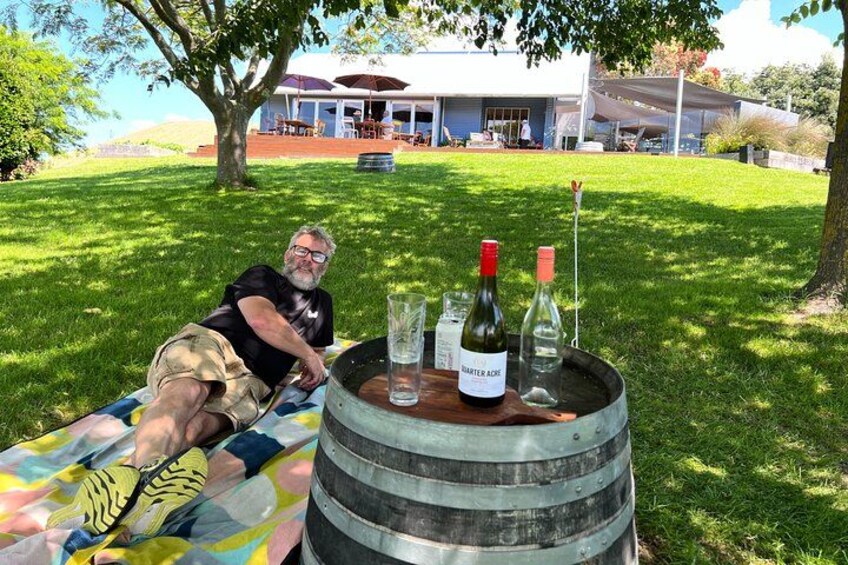 A man sitting on a colorful blanket outdoors with a wooden barrel containing wine bottles and glasses. 1