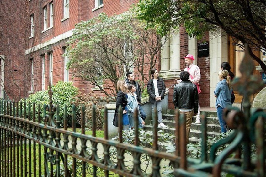 A group of people standing on steps in front of a red brick building with a tree and a metal fence in the foreground. 2