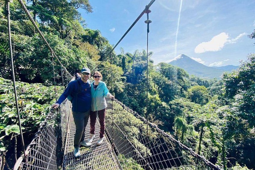Two people on a suspension bridge in a forested area with a mountain in the background. 1