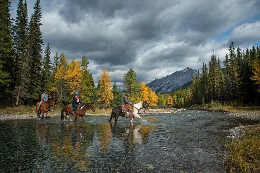 Trois personnes à cheval, se promenant le long d’une rivière, avec des arbres aux couleurs automnales et des montagnes en arrière-plan. 2