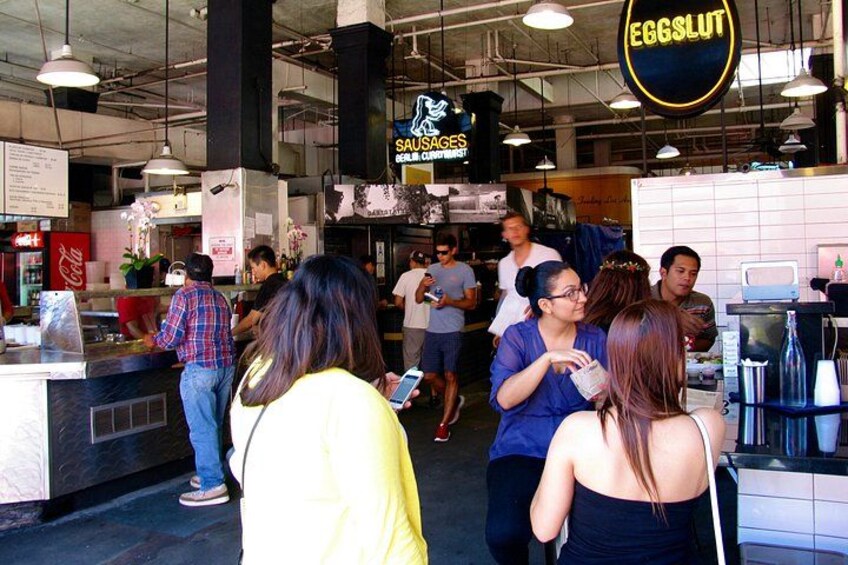 A busy food court with people dining and a sign for 'SAUSAGES'. 2
