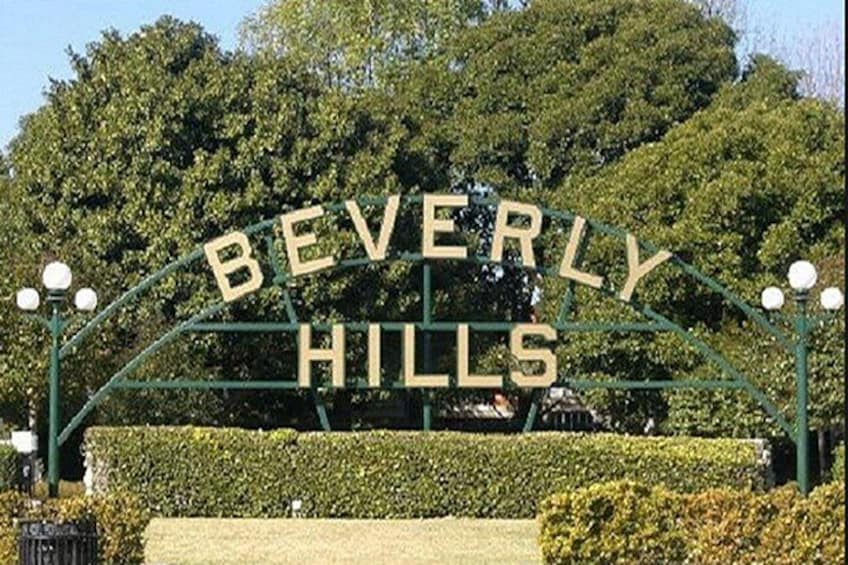 Un cartello con la scritta 'BEVERLY HILLS', sormontato da un arco verde e caratterizzato da scritte bianche, circondato da siepi e alberi curati. 1