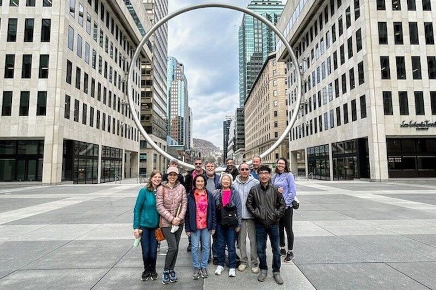 En grupp människor som står på ett torg med moderna byggnader och en stor, cirkelformad konstruktion i bakgrunden. 1