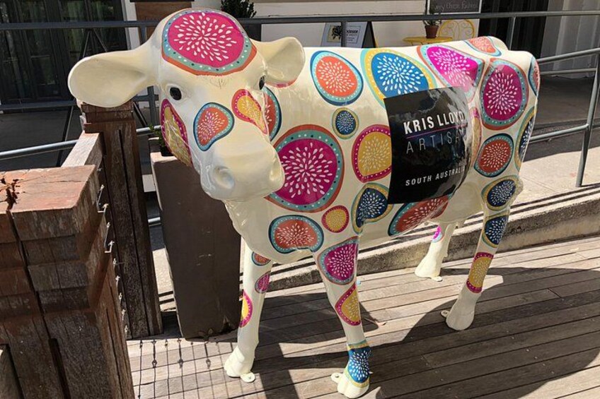 A colorful cow sculpture with a label reading 'Kris Lloyd Artis' on a wooden platform. 3
