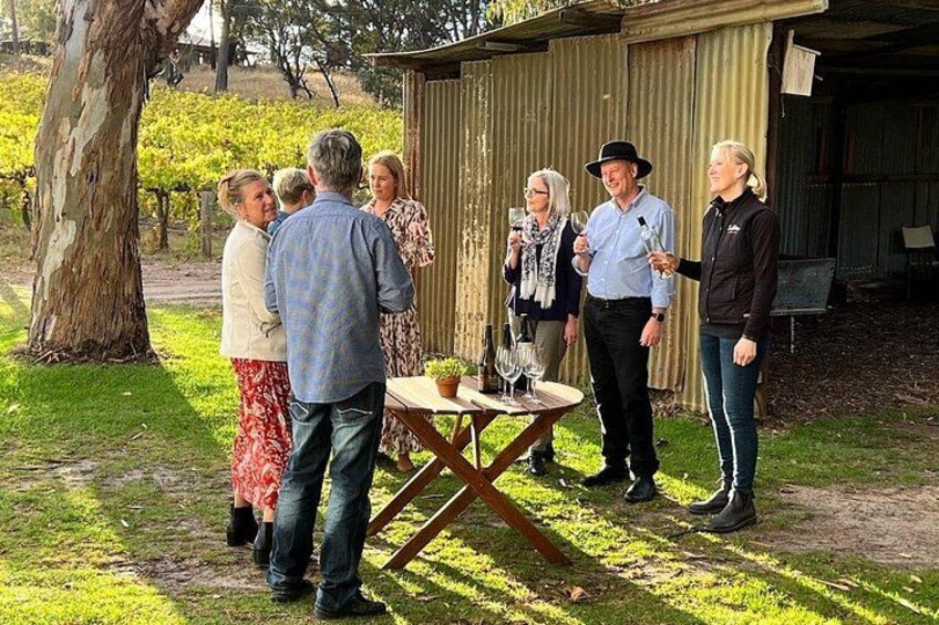 En gruppe mennesker står udendørs nær et bord med vinglas og flasker, i en vinmarkssetting. 5