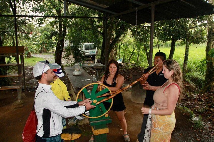 Un grupo de personas interactuando con una gran rueda de madera al aire libre, rodeado de árboles y una estructura cubierta. 3
