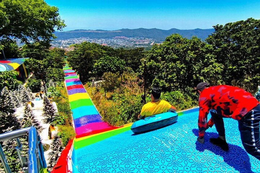 A person sliding down a rainbow-colored slide, another person assisting, and a scenic view of trees and mountains in the background. 5
