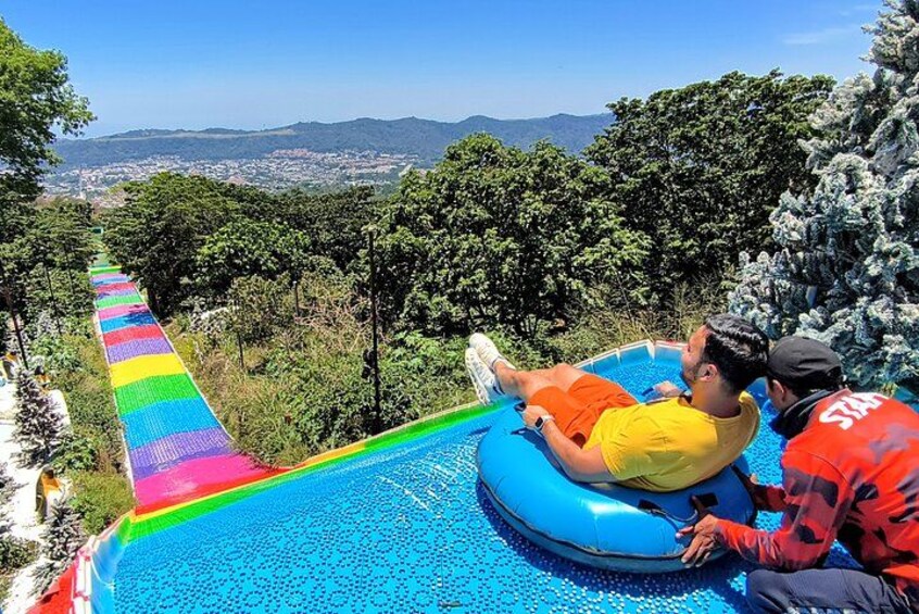 A person lying on an inflatable tube on a blue water slide with a rainbow-colored path, surrounded by trees and a view of a distant landscape. 1
