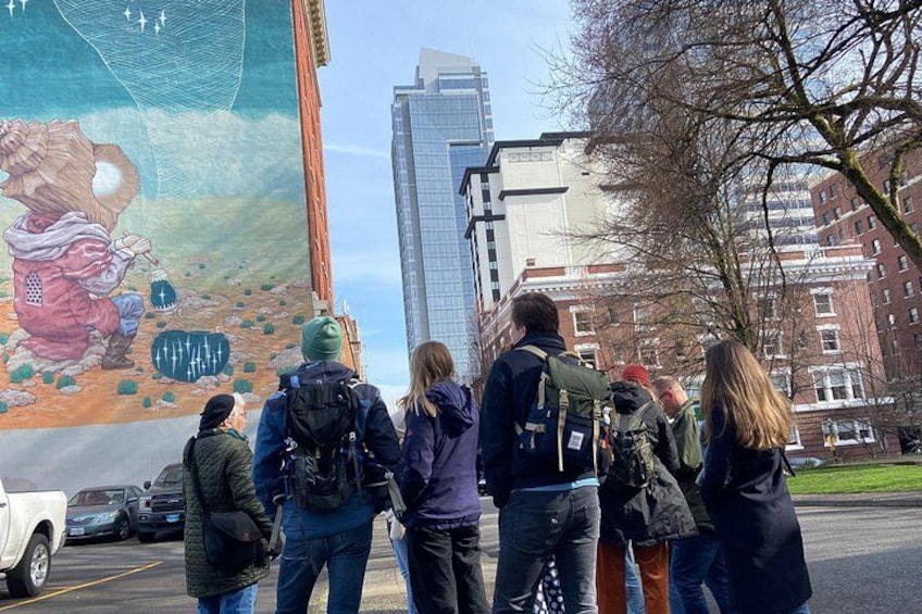 Un groupe de personnes se tient sur un parking, observant une grande fresque murale sur un bâtiment. 4