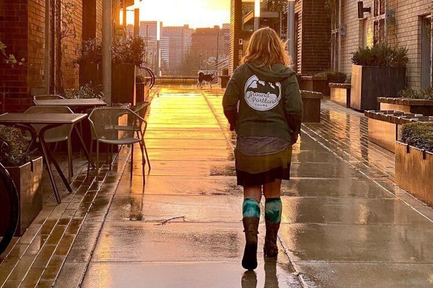 A person walking on a wet sidewalk with tables and chairs on the left, buildings on both sides, and a sunset in the background. 4