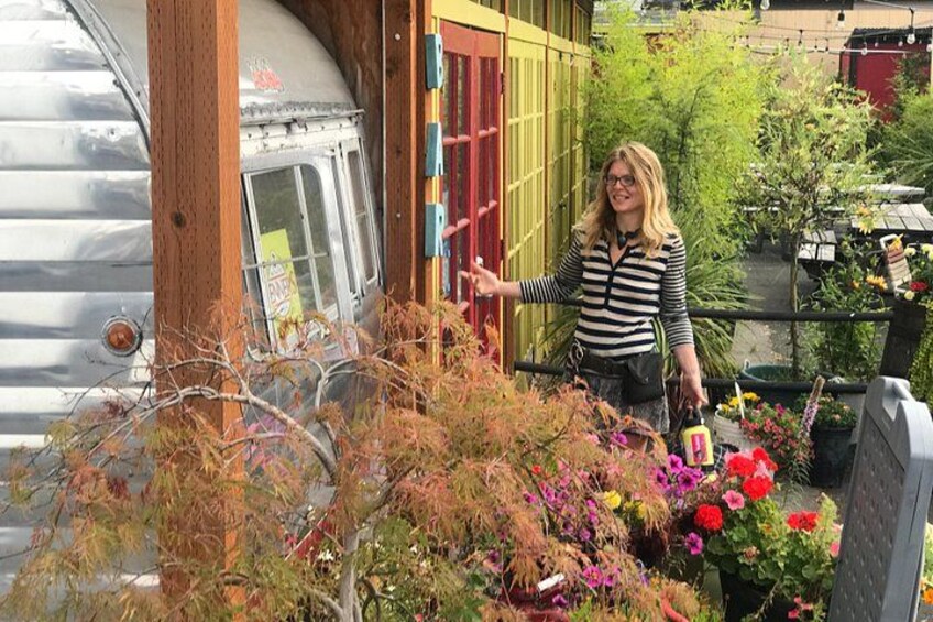 Una persona de pie junto a una caravana plateada antigua con puerta de madera, rodeada de diversas plantas y flores. 2