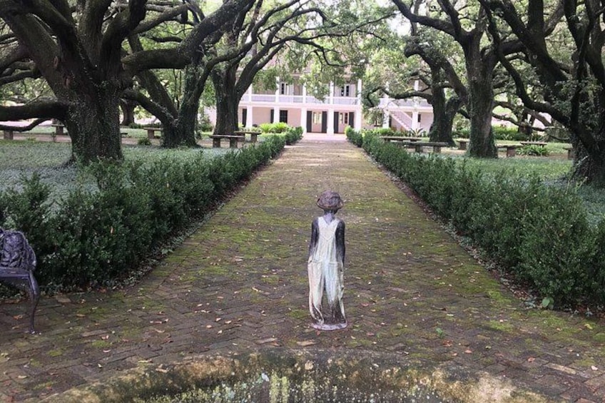 A statue of a woman in a long dress on a paved path flanked by trees and a building in the background. 1