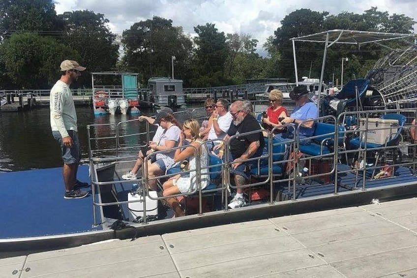 Een groep mensen op een boot aan een steiger, met water en bomen op de achtergrond. 3