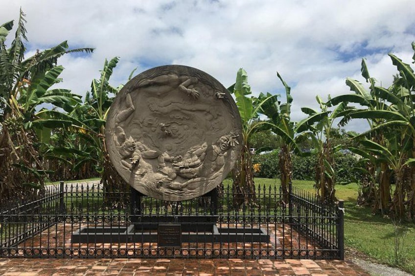 A large spherical sculpture with intricate carvings, surrounded by a black metal fence, set in a garden with banana trees. 1