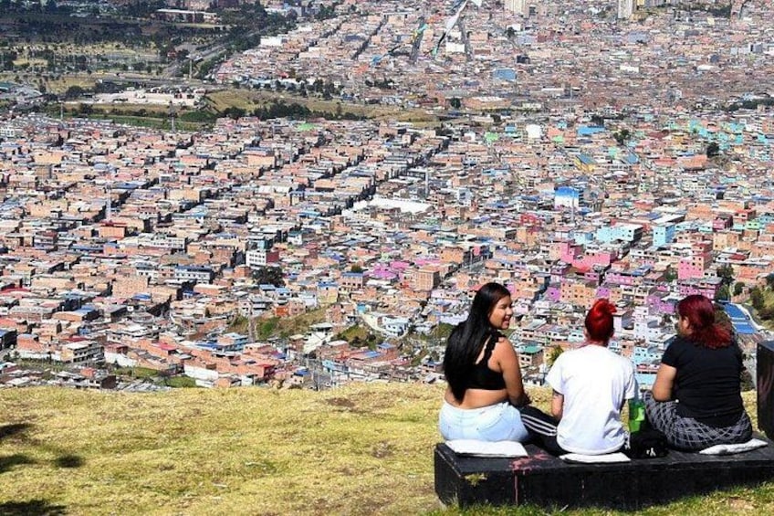 Three women sitting on a bench overlooking a densely packed urban landscape. 4