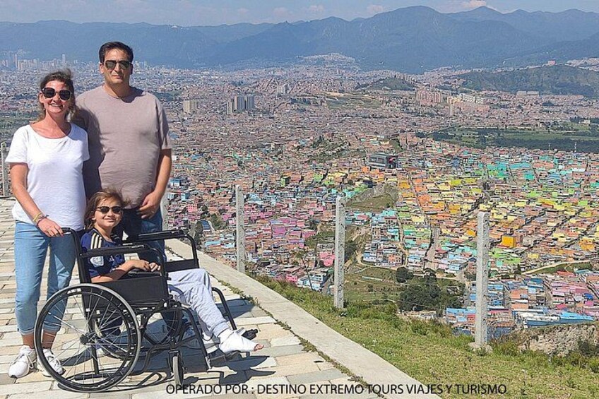 A family with a child in a wheelchair, standing on a viewing platform overlooking a densely populated cityscape with colorful buildings. 4