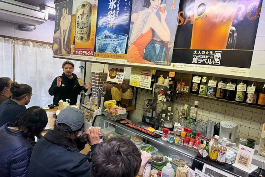 A bar with people seated at tables, a bartender, and various bottles and condiments on shelves. 3