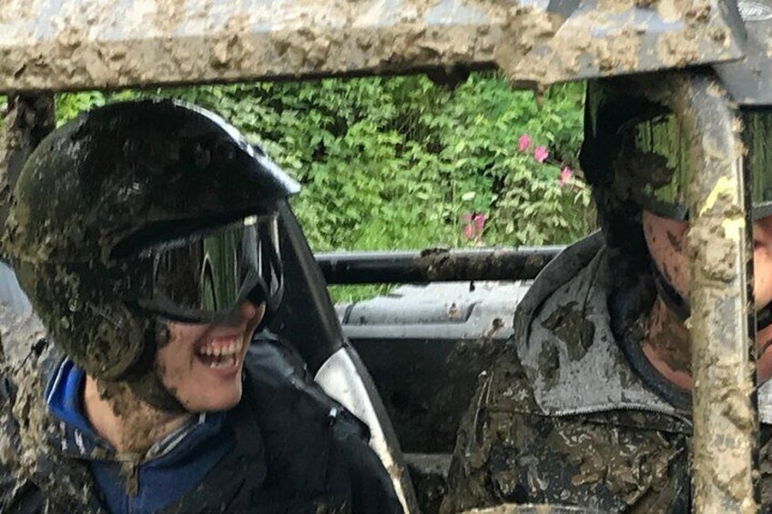 Two people in a vehicle covered in mud, wearing helmets and protective gear. 1