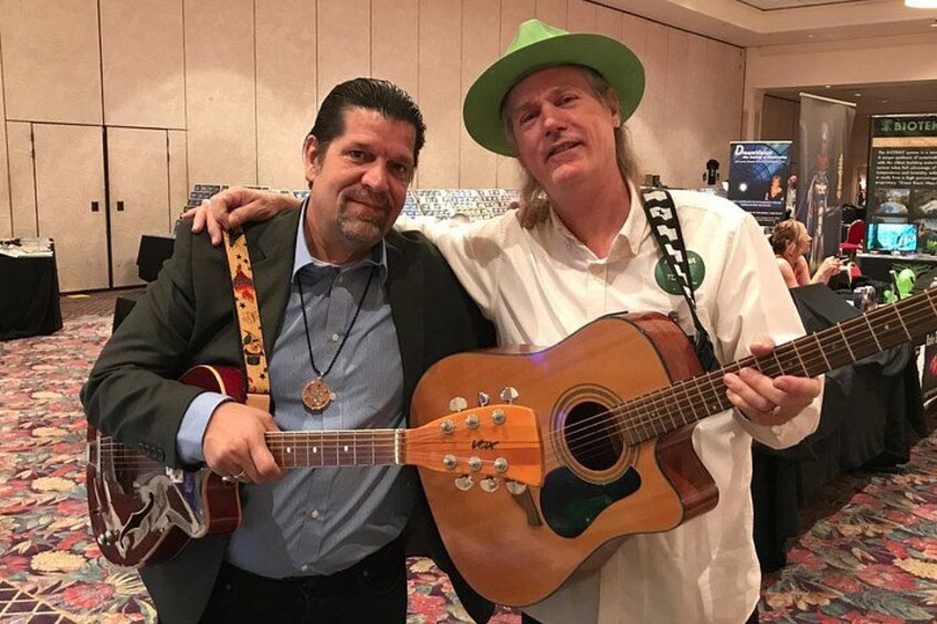 Dos personas sosteniendo guitarras en un salón de conferencias con una alfombra floral y un cartel de fondo. 4