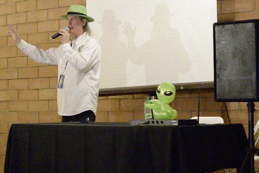 Una persona hablando en un micrófono, una figura alienígena verde y una pantalla de proyección con la sombra de alguien. 3