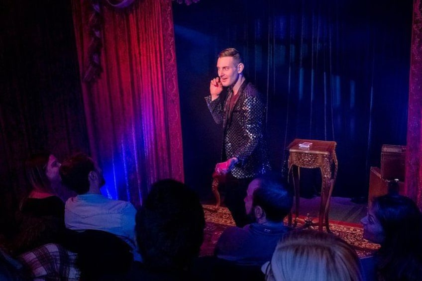 A person on stage with a microphone, audience seated in front, and a small table on stage. 2