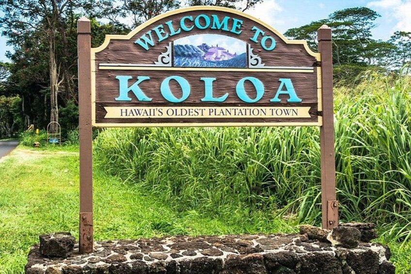 A wooden welcome sign for Koloa, Hawaii's oldest plantation town, with a stone base and lush greenery in the background. 2
