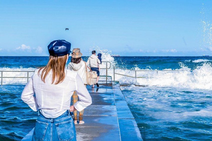 A pier extending into the ocean with people standing at the end, waves crashing against the pier, and a clear blue sky. 3
