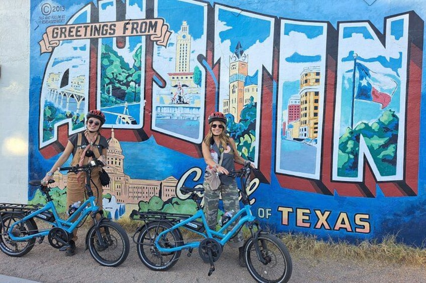 En väggmålning med två personer på cyklar, stora bokstäverna A och T, och texten ”Hälsningar från Austin, Texas”. 1