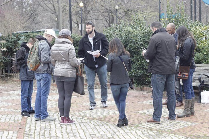 Un grupo de personas de pie sobre una superficie pavimentada, algunas sosteniendo documentos, en un entorno exterior con árboles y un banco al fondo. 3
