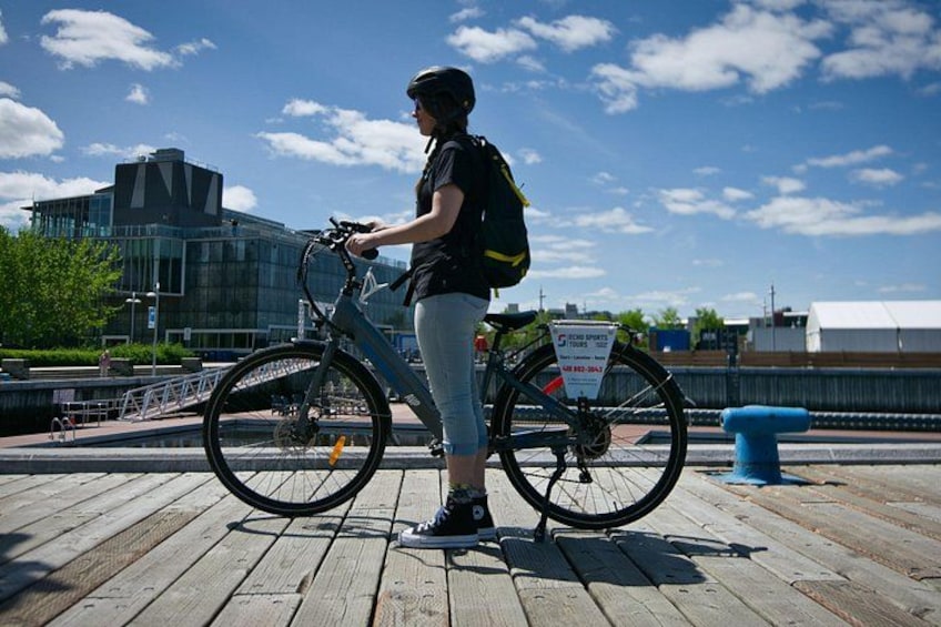 Eine Person steht neben einem Fahrrad auf einer Holzpromenade, im Hintergrund ein modernes Gebäude. 3