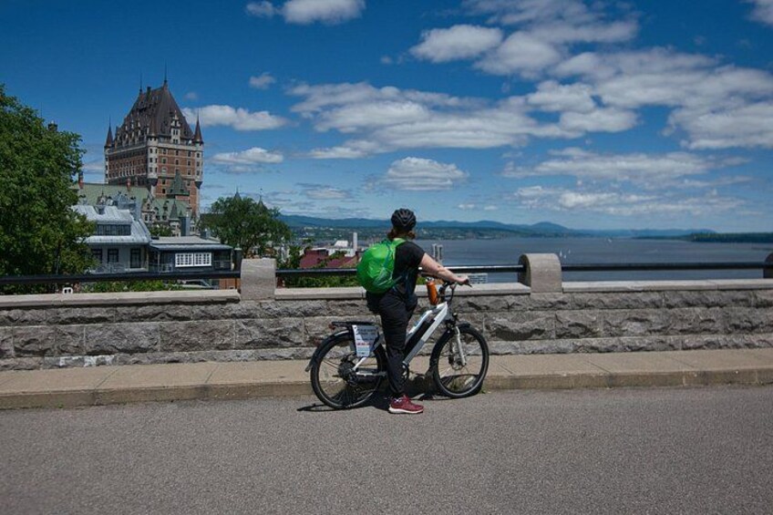 A person standing next to a bicycle with a green backpack, overlooking a body of water and a historic building. 2