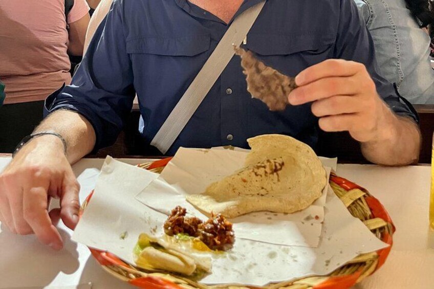 A person holding a piece of flatbread, with a basket containing a wrapped food item and some vegetables on a table. 4