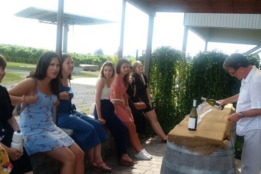 A group of people sitting outdoors, a man pouring wine from a bottle, and a table with a bottle and glasses. 1