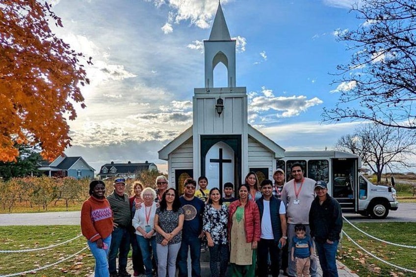 Un groupe de personnes se tient devant une église, avec une croix, un véhicule et des arbres aux couleurs automnales. 2