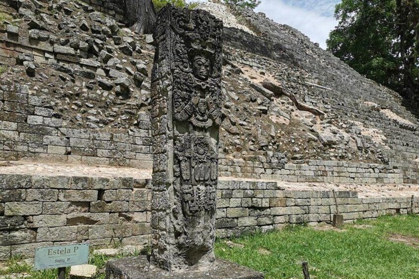 A stone structure with carvings, a sign labeled 'Estela P,' and a grassy area. 1