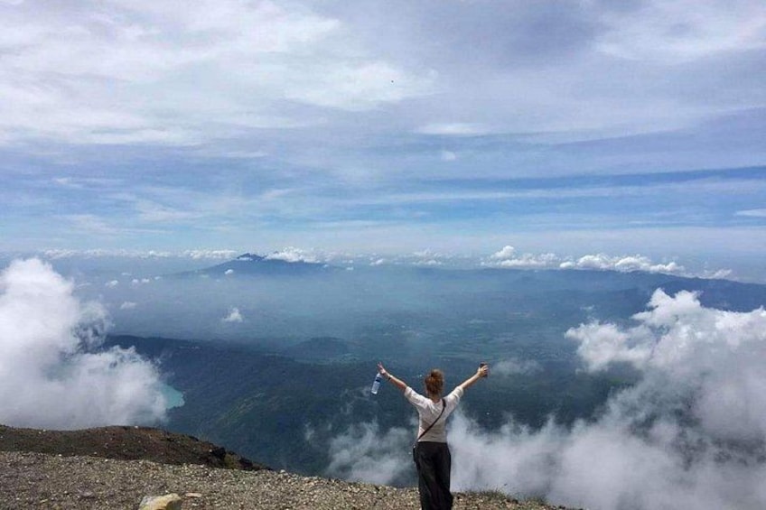 Una persona de pie sobre un afloramiento rocoso con los brazos levantados, contemplando un extenso paisaje de nubes y montañas lejanas. 2
