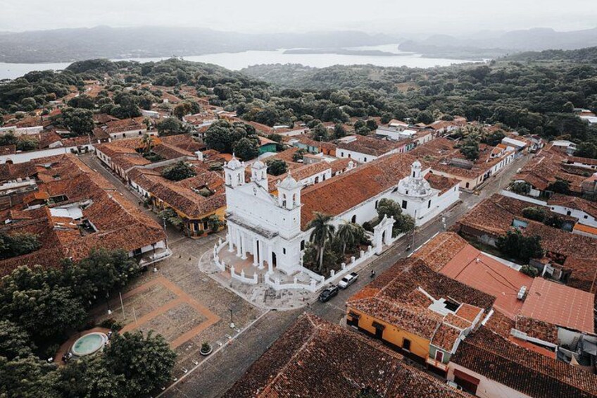 An aerial view of a historic town with a prominent white church, terracotta-roofed buildings, and a large body of water in the background. 1