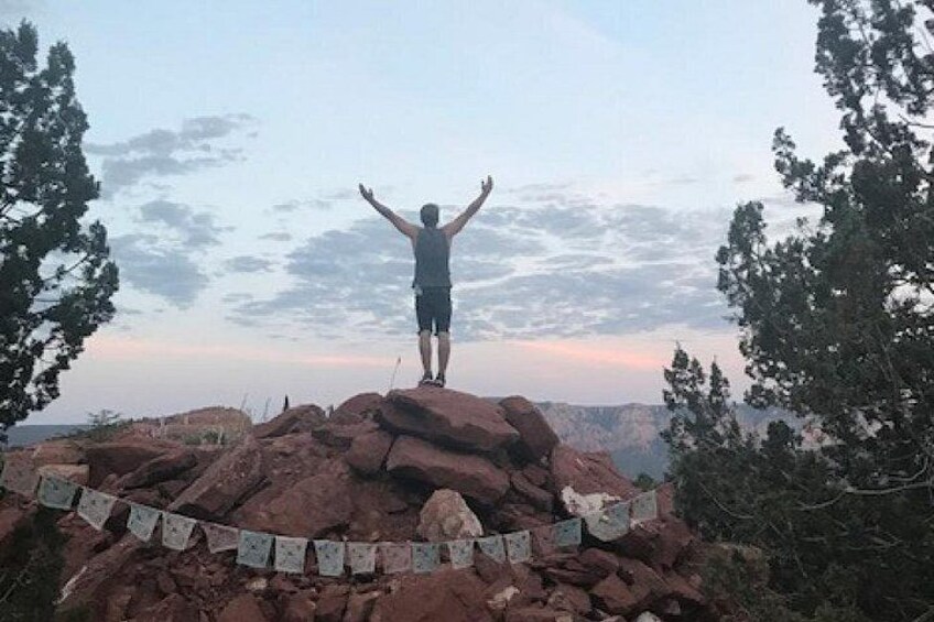 A person standing on a rock formation with arms raised, surrounded by trees and a clear sky. 2